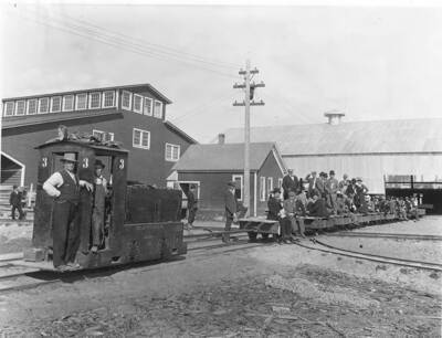 According to the writing on the back of the photograph, these are Japanese commissioners that have come to a mill to see the wood products being produced. Most of them are sitting on the flat railcars. In the middle of the group of Japanese commissioners are two women. Two men stand in the engine to the left-hand side of the photograph.
