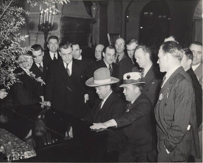 Men gather around a piano and are singing while two men play.