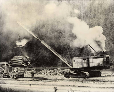 A large crane loads a large bundle of logs on to the back of a trailer while a man watches. Behind them, are more logs waiting to be loaded for transport. Same photo can be found in Box 203 Folder 42 with the following description: 'The description on the back of the photograph reads 'The monster (also named Big Bertha): Old coulee Dam Irrigation Project Drag Bucket. Converted to log unloading and decking machine at log drive landing on N. fk. Clearwater just above Little NF. Hangled entire truck load of logs.'