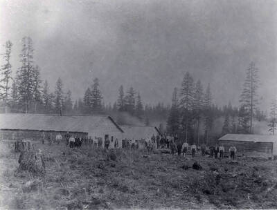 Men pose for a picture in front of buildings at an early logging in Latah county, Idaho. The area in front of the men is cleared of trees while the area behind the buildings is thick with trees.