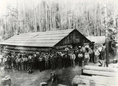 A group of loggers (and a dog) stand in front of building in an unknown logging camp near Bovill, Idaho. Behind the men and buildings are un-harvested trees, while in the lower right hand section of the photograph are harvested logs.