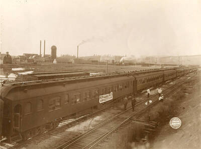 Several locomotive cards sit while six men stand in front of them. There is a sign on one of the train cars that partially reads 'Spokane Wash September 24-27 1913'.