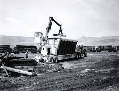 A man watches as a log is lowered into a "smokeless trash burner." Description taken from envelope holding the photographs. In the background are railcars full of logs wait.