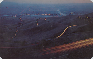 Postcard is a photograph of the old spiral highway above Lewiston, Idaho at dusk.  | Lewiston Hill at dusk. In this time exposure, each car lights its tortuous course over the ten mile route against a back-drop of the lights of Lewiston, Ida. and Clarkston, Wash.
