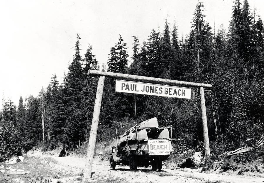 A canoe-laden automobile passing beneath a sign that reads 'Paul Jones Beach.' Priest Lake, Idaho. Donated by Marjorie (Paul) Roberts through Priest Lake Museum.