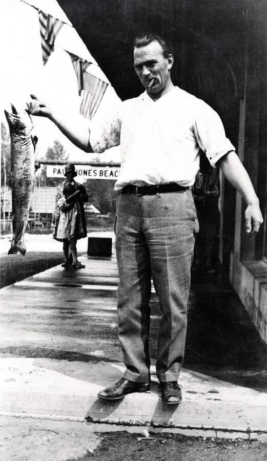 Leonard Paul posing with a fish he caught. He is smoking a cigar. A woman and young girl stand in the background. Coolin, Idaho. Donated by Marjorie (Paul) Roberts through Priest Lake Museum.