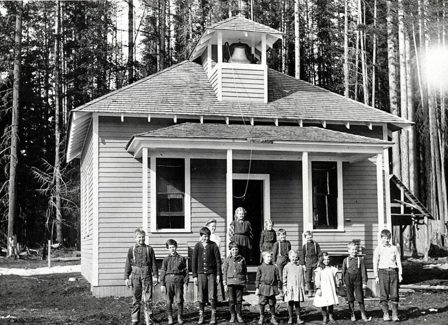 Thirteen children and a teacher pose in front of a school building.