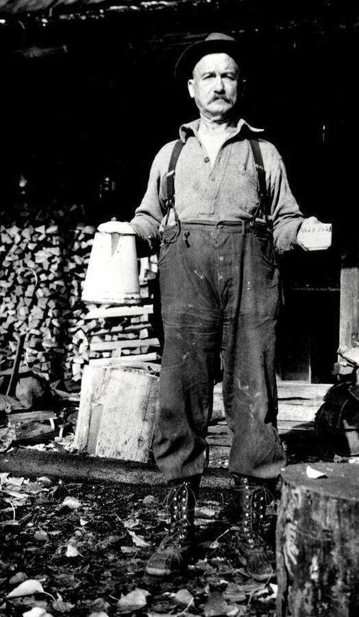 Frank Brown standing in front of Dad Moulton's cabin, holding a kettle and cup.