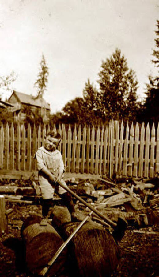 Lee Calfee trying to hold an axe next to a woodpile. Donated by Margaret Randall through Priest Lake Museum.