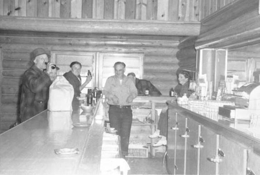Interior view of Nordman Bar. Six people stand around the bar. Donated by Mike Winslow through Priest Lake Museum.