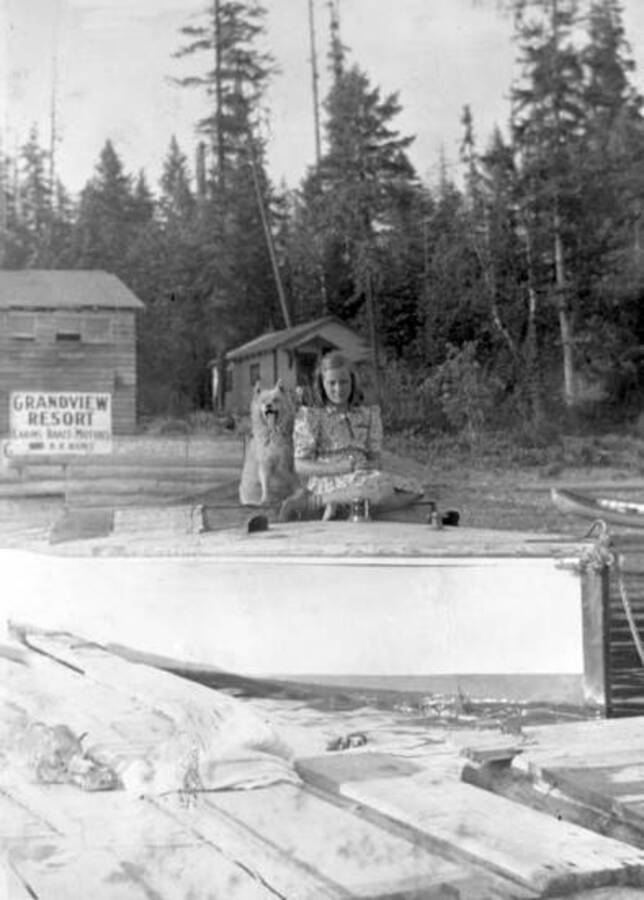 A girl with a dog sitting in a boat docked at Grandview Lodge. Priest Lake, Idaho. Donated by Mary Hunt through Priest Lake Museum.