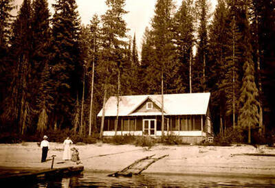 Three people on the beach in front of a cabin on the lake (Charles Schrimpf?). Donated by Viv Beardmore through Priest Lake Museum.