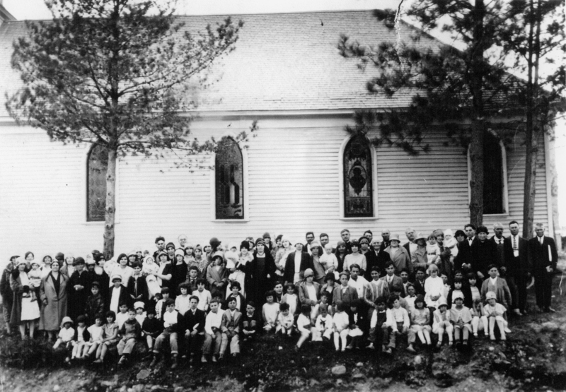 A large group of people gathered in front of a building with arched windows, possibly a church, with trees on either side. The group includes adults and children, dressed in different styles of clothing.