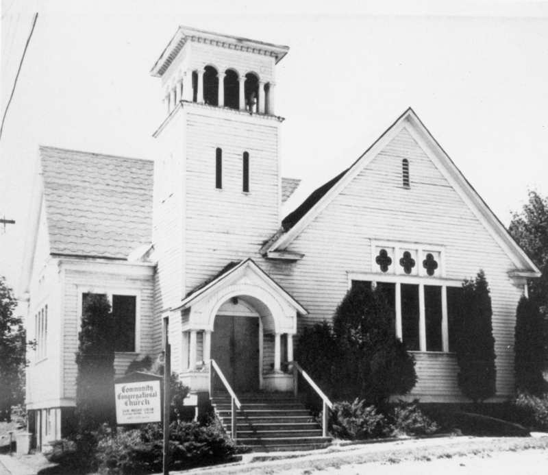 A church building with a tall bell tower featuring arched openings. The entrance has a small roofed porch with steps leading up to double doors. There are large windows with decorative designs above them. A sign in front of the church reads "Community Congregational Church." Shrubs and trees are planted around the building. The sign near the entrance reads: "Community Congregational Church... Church School... 10:00 A.M. Worship 11:00 A.M."