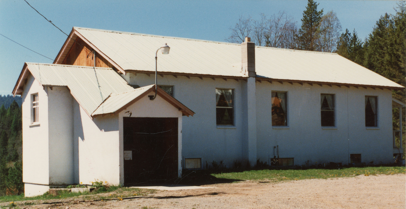 A single-story building with a white exterior and a metal roof, surrounded by trees. It has several windows and a small entrance with a light fixture above it. A chimney extends from the roof on one side, and there is a grassy area in front.