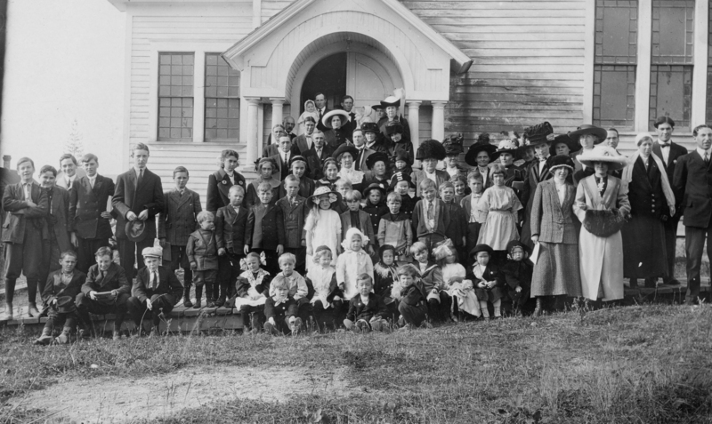 A large group of people, including men, women, and children, stand in front of a building with a porch and arched doorway. The adults are mostly wearing formal clothing and hats, while the children are dressed in a variety of suits and dresses. Some children sit on the ground, while others stand or are held by adults. The building has large windows with decorative panes.