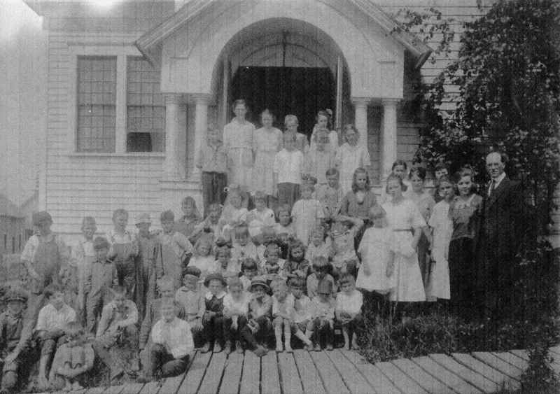 A large group of children and a few adults standing on the steps of a building with a wooden walkway leading up to it. The building has an arched entrance and large windows. Some children are seated, while others stand further up the steps, with one man standing to the right of the group.