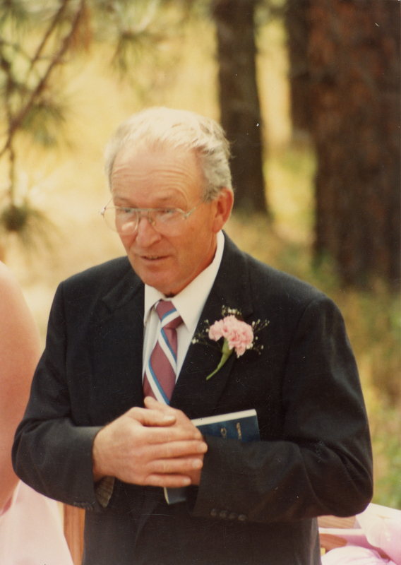 An older man wearing a suit and tie stands outside, holding a book. He has glasses and a pink flower pinned to his jacket. Trees are visible in the background.