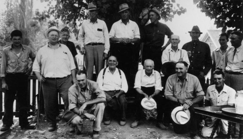A group of men, mostly in shirts and hats, are gathered outdoors. Some are standing while others are seated in front. They are under a tree with a fence and buildings in the background. A table is visible to one side.