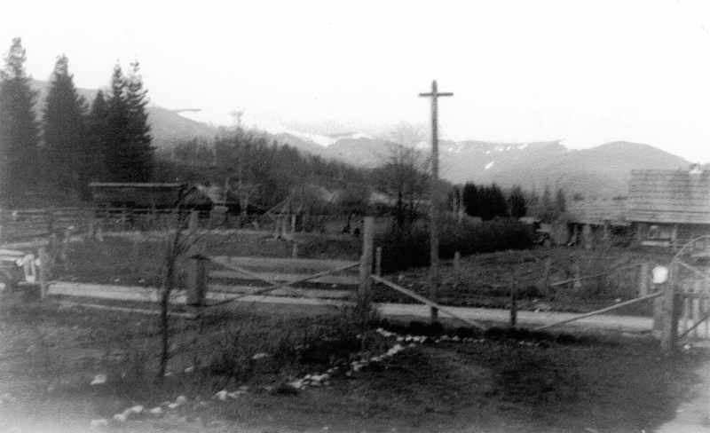 A rural scene with a wooden fence and gate in the foreground, a large cross on a pole, and several trees. There are hills and a small structure in the background.