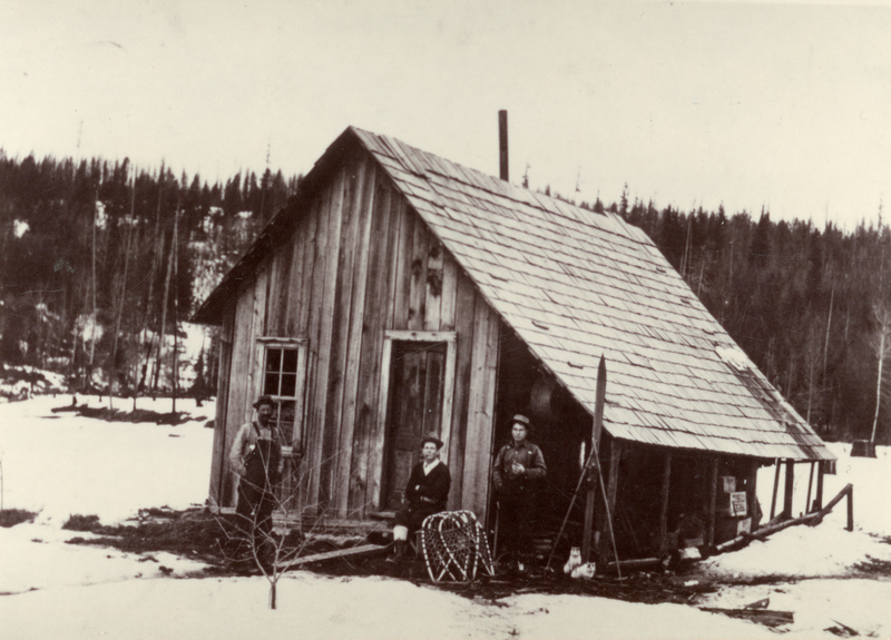 Three people standing outside a wooden cabin in a snowy landscape with trees in the background. One person is leaning against the cabin, another is sitting on the porch, and the third is holding skis and snowshoes are lying nearby.