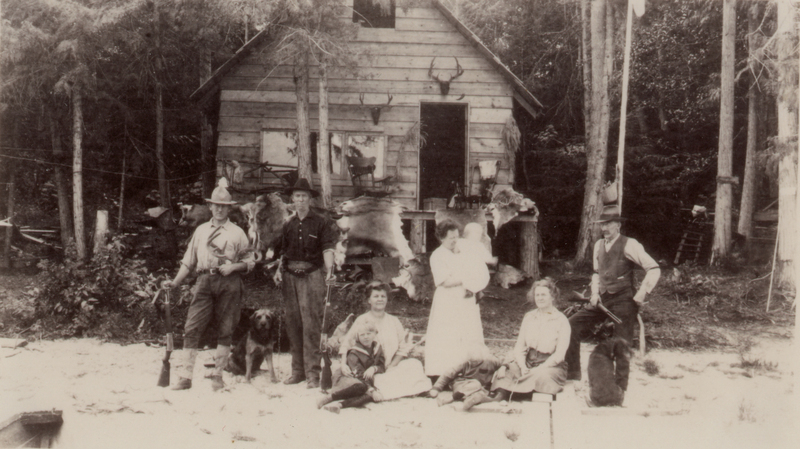 A group of seven people, including men, women, and children, are posed outside a wooden cabin. Two men are standing with rifles, accompanied by two dogs. One woman is holding a baby, while another two women and two children are sitting on the ground. The cabin has animal skins hanging and antlers mounted above the door, and is surrounded by trees.