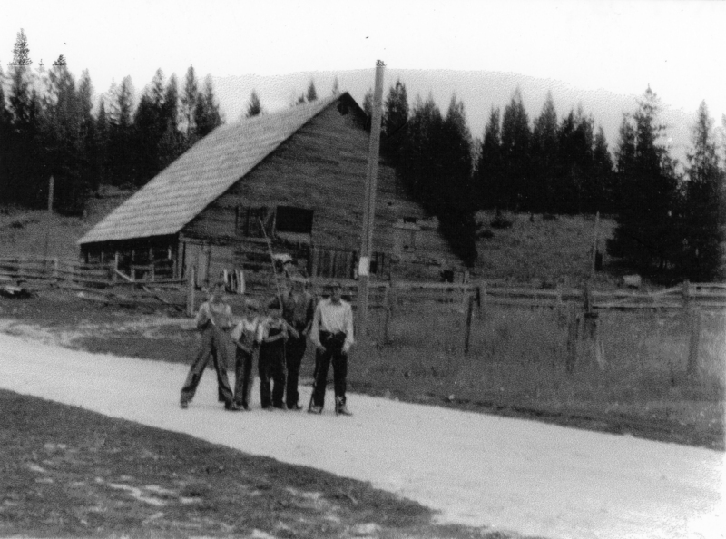A group of five people standing on a road in front of a wooden barn, surrounded by a fenced area and trees in the background.
