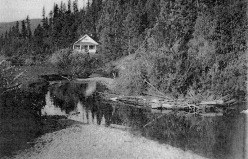 A small house with a porch is situated near a forested area. In the foreground, there is a calm body of water reflecting the surrounding trees and bushes.