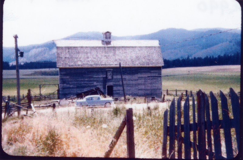 A wooden barn with a small cupola on top situated in a rural landscape with mountains in the background. A vintage car is parked in front of the barn, surrounded by a fence and a field with tall grass in the foreground. A utility pole is also visible near the barn.