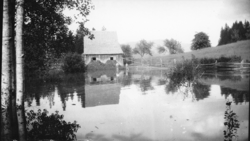 A wooden house with a steep roof is partially surrounded by water, reflecting its image. Two people stand in shallow water near a wooden fence. Trees and bushes are visible around the scene.
