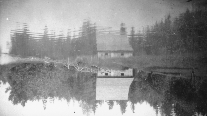 A reflection of a rural scene on water, showing a house with a sloped roof, surrounded by trees and vegetation. The water has slight ripples, distorting the reflection.