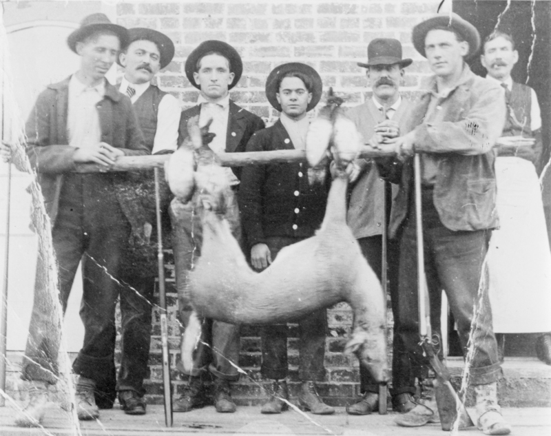 A group of eight men standing in a line in front of a brick wall. They are holding a hunting rifle and an animal hanging upside down from a wooden pole. The men are wearing hats and formal or work attire.