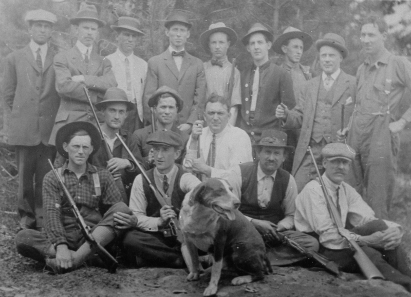 A group of sixteen men and one dog. The men are dressed formally, with some wearing hats and suits. Many of them are holding rifles. The dog is sitting in the front near the center. The setting appears outdoors, possibly in a forested area.