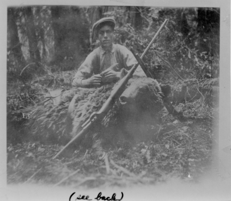 A person wearing a hat is sitting in a forested area with a large bear lying in front of them. A rifle is resting on the bear. Text on photo reads "See Back".