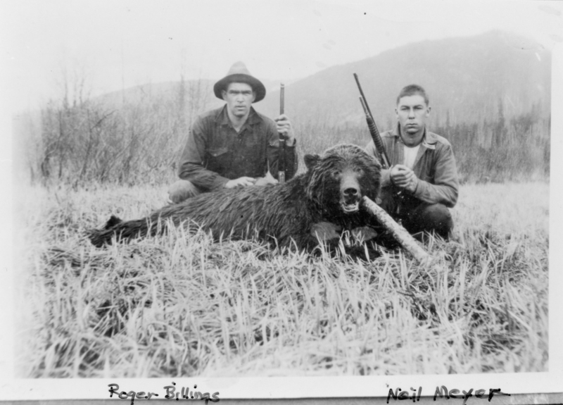 Two men are crouching in a grassy field, each holding a rifle. In front of them lies a large bear with its mouth open. There are mountains and trees in the background. The text at the bottom of the image reads "Roger Billings" on the left and "Neil Meyer" on the right.