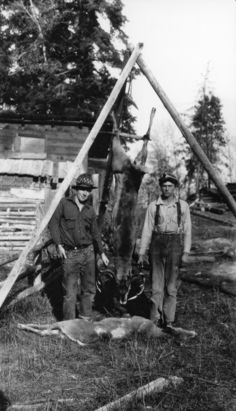 Two men stand next to a wooden A-frame structure outdoors. A deer is hanging upside down from the structure, and another deer lies on the ground in front of them. A rustic building and trees are in the background.