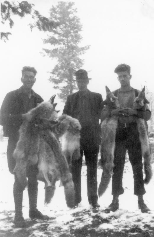 Three men stand outdoors, each holding animal pelts. The background shows a large tree, and the ground is covered with snow. Two of the men wear hats, and all are dressed in jackets and boots.