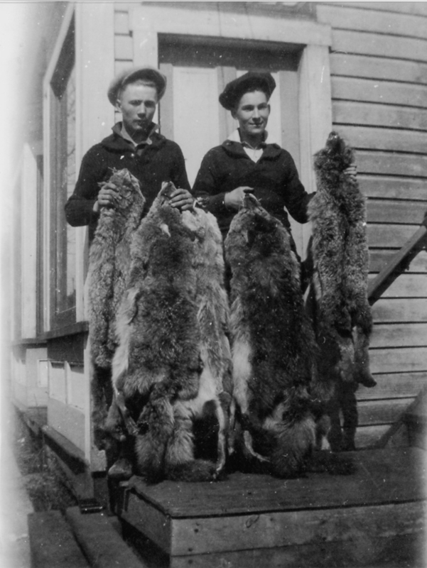 Two people standing on steps outside a building, each holding several animal pelts. They are dressed in dark clothing and hats. The building has wooden siding and a visible doorway behind them.