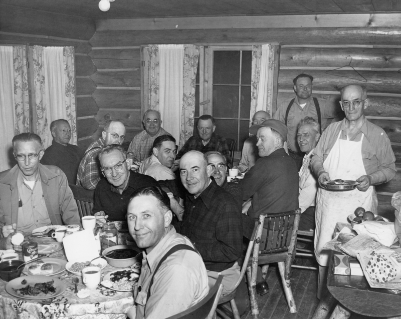 A group of men gathered around a table inside a log cabin. The table is covered with dishes, food, and cups. Some men are seated, while others stand or lean against the wall. One man wearing an apron holds a tray. The cabin has wooden walls and is decorated with curtains.