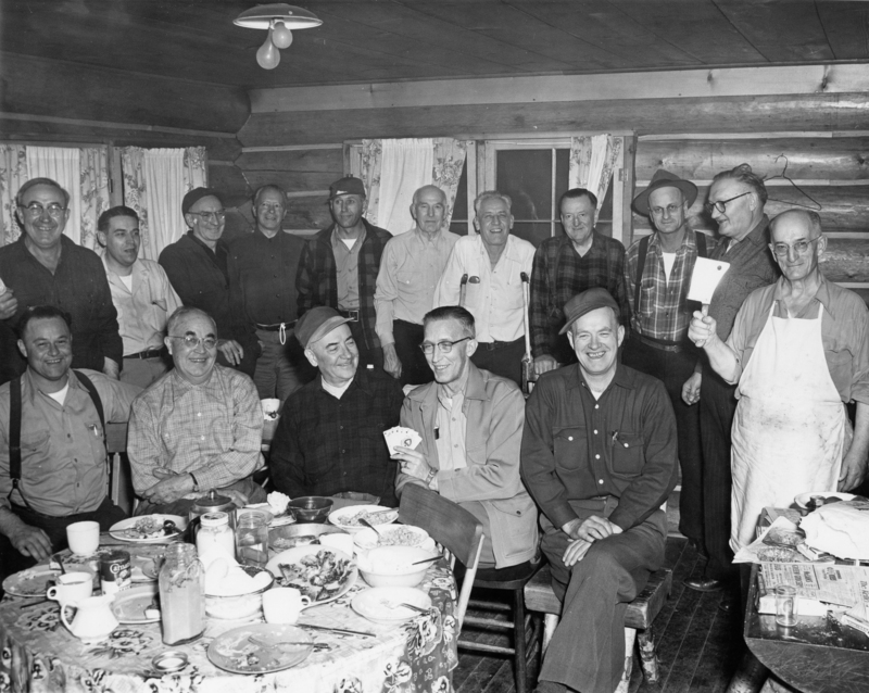 A group of men gathered in a log cabin, with some sitting around a table covered in dishes and food, while others stand behind them. One man is holding playing cards, and another is holding a meat cleaver. The men wear casual, rugged clothing.