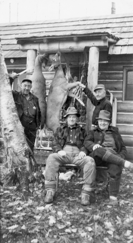 Four men are posing with two hanging deer outside a rustic cabin. Two men are standing next to the deer, and two are seated in front of them on chairs. The ground is covered with fallen leaves.