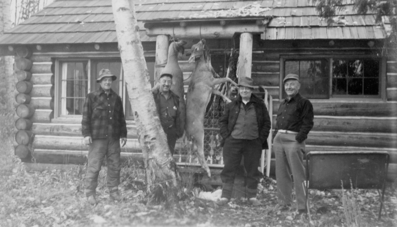 Four men stand in front of a log cabin. Two deer are hanging from a wooden structure next to them. The ground is covered in leaves and there is a birch tree at the forefront. The men are dressed in outdoor clothing and hats.