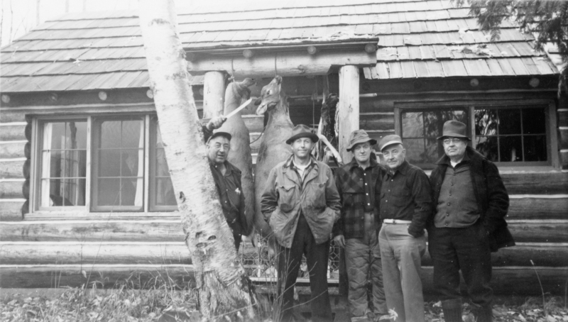Five men standing in front of a log cabin, with two deer hanging behind them. A tree trunk is visible in the foreground. The men are dressed in outdoor clothing, including hats and jackets.