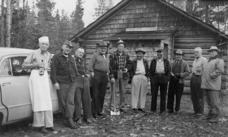 A group of men standing in a row outside a wooden building. One man is leaning against a car holding a bottle, wearing an apron and hat. The others are dressed in a mix of casual and work clothes, some wearing hats. A wooded area is visible in the background.