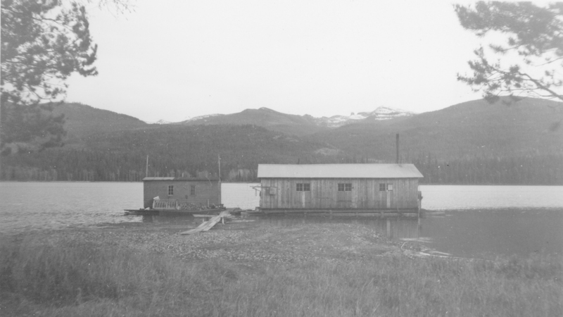 Two wooden buildings float on a body of water, surrounded by a mountainous landscape with trees. One building is larger, with a simple roof and visible windows. The shoreline is grassy, and trees frame the scene on either side.