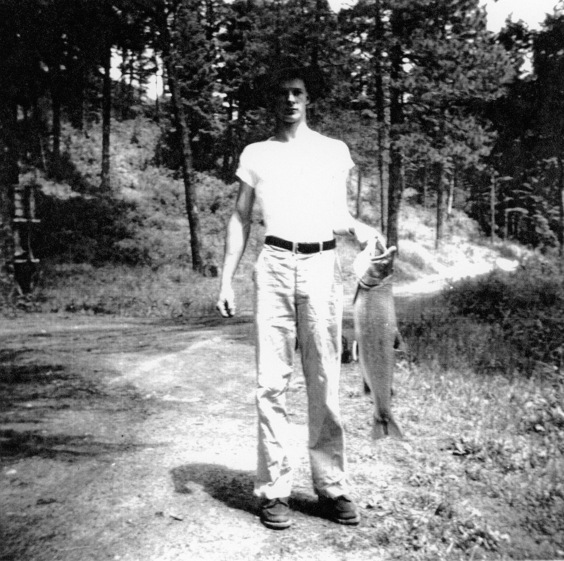 A person standing on a dirt path in a forested area, holding a large fish in one hand.