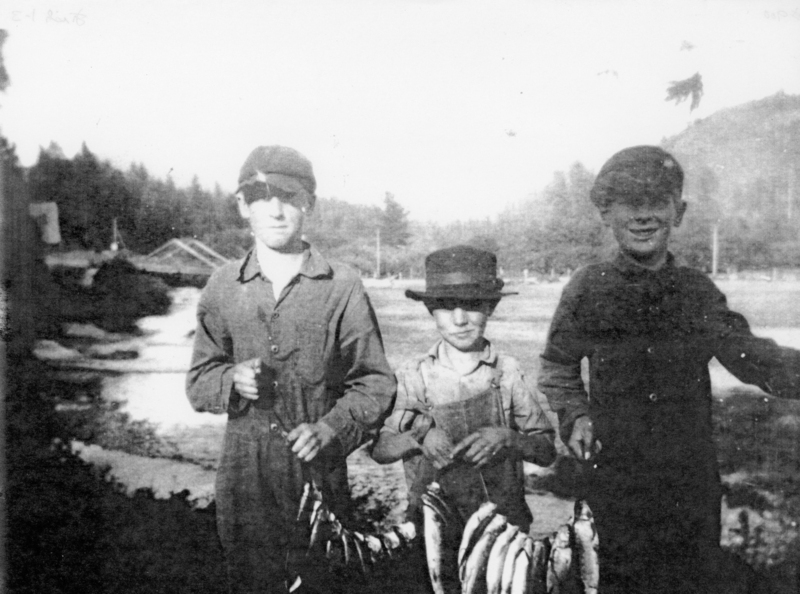 Three young boys stand outdoors, each holding strings of several fish. A body of water and a wooden structure are visible in the background.