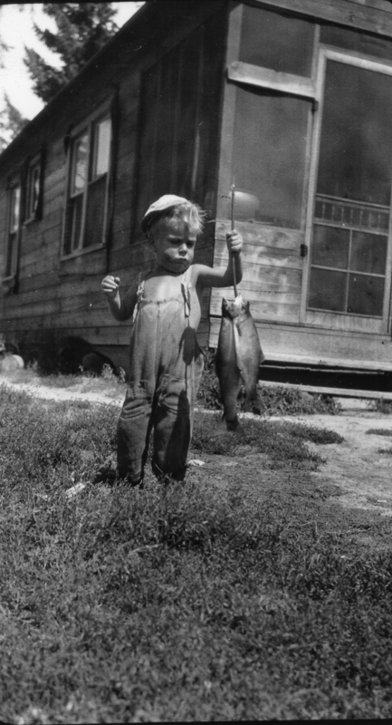 A small child standing on grass in front of a wooden house, holding a large fish by a string or pole. The child is wearing overalls and a cap.