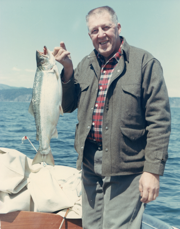 A man standing on a boat holding a large fish, with a body of water and distant hills in the background. He is wearing a plaid shirt under a jacket.