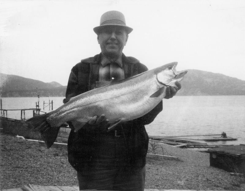 A person wearing a hat and jacket holds a large fish in front of them. The background shows a lakeside setting with mountains and a dock area.